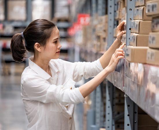 Smart Asian young woman happy to work in store warehouse. Girl s Smart Asian young woman happy to work in store warehouse. Girl standing and checking quality and quantity of inventory stock on shelf by model name and serial number it show on packaging of goods.