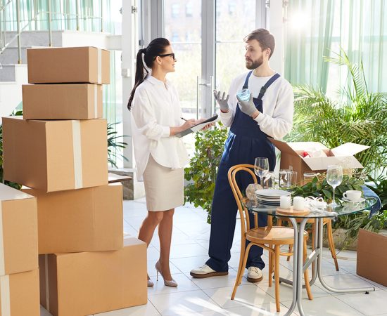 Content confident female event manager with pony tail making notes in paper while holding clipboard and discussing goods replacement with mover while preparing space for event
